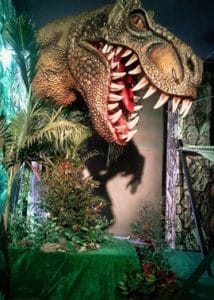 Escultura monumental de la cabeza abierta de un T-Rex ambientada con vegetación simulada en un espacio de exhibición, fabricada mediante impresión 3D por CR Studio 3D en Medellín.