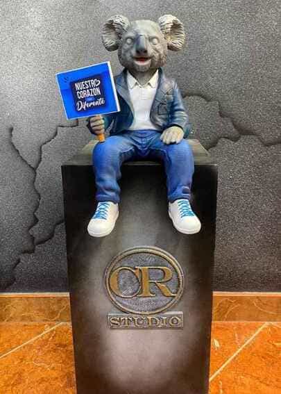 Escultura corporativa de la mascota de Qualá (koala) vestida con chaqueta y jeans sosteniendo un letrero de "Nuestro Corazón es Diferente", fabricada mediante impresión 3D por CR Studio 3D en Medellín.